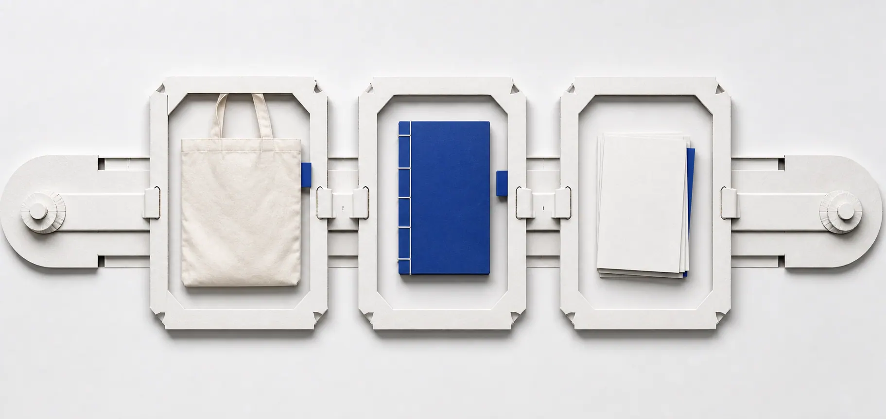 A blank canvas tote bag, thread-bound notebook and postcard stack moving through a silver paper sample track